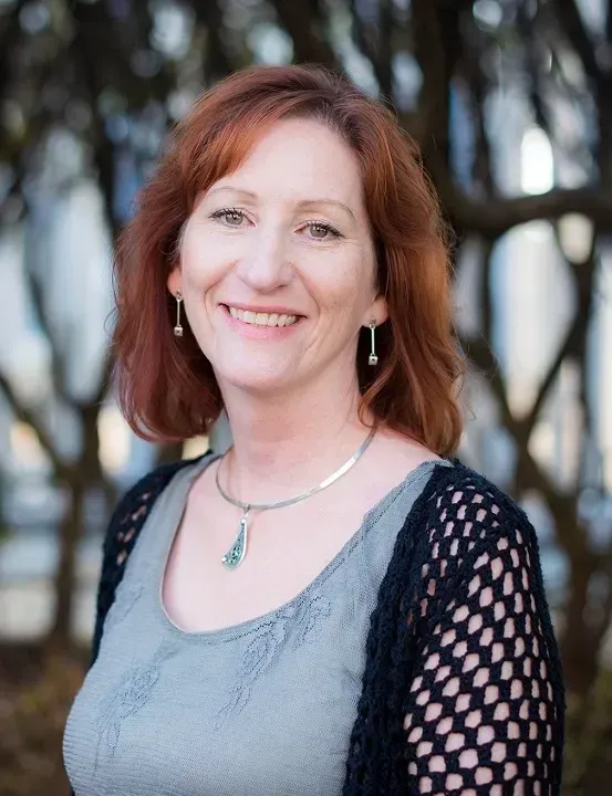 A woman with red hair is smiling and wearing a necklace and earrings.