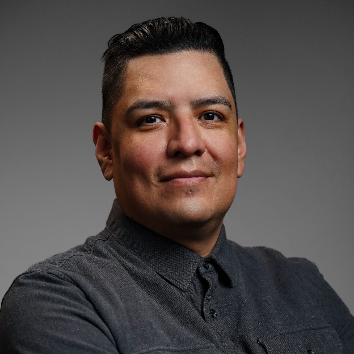 A man in a gray shirt is smiling for the camera with his arms crossed.