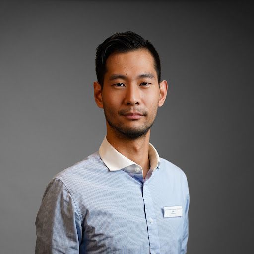 A man with a beard is wearing a blue shirt and standing in front of a gray background.