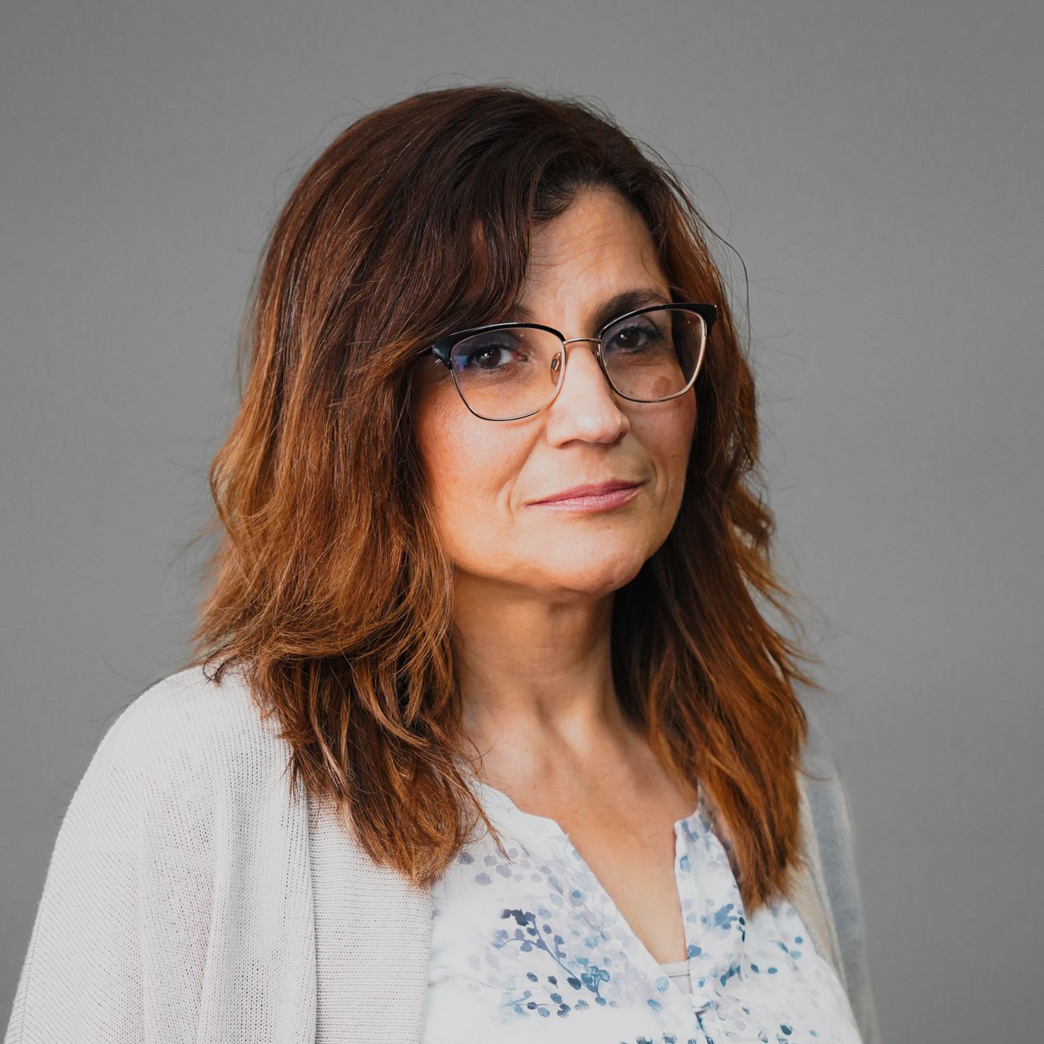 A woman wearing glasses and a sweater is standing in front of a gray background.