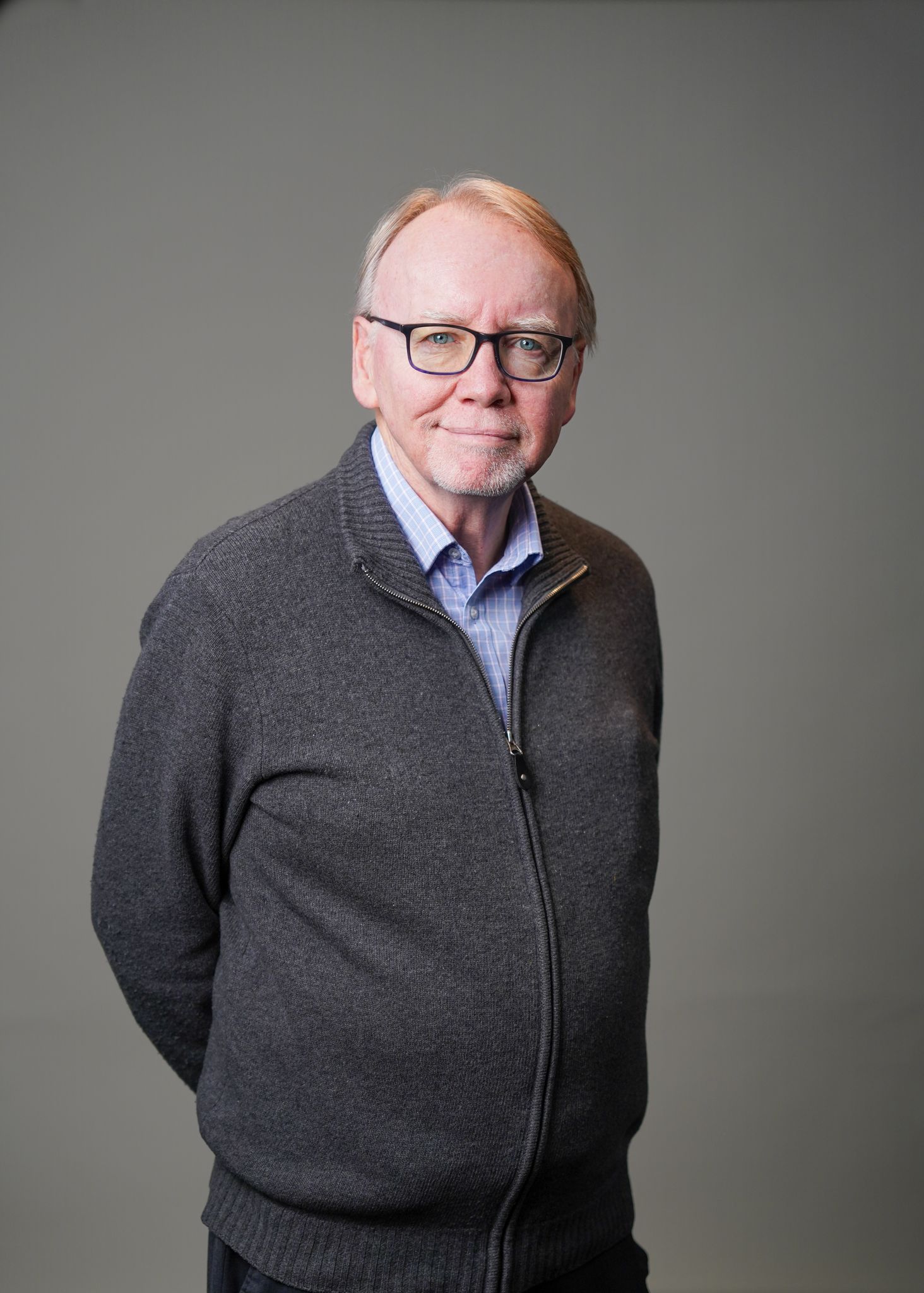 A man wearing glasses and a grey sweater is standing with his arms behind his back.