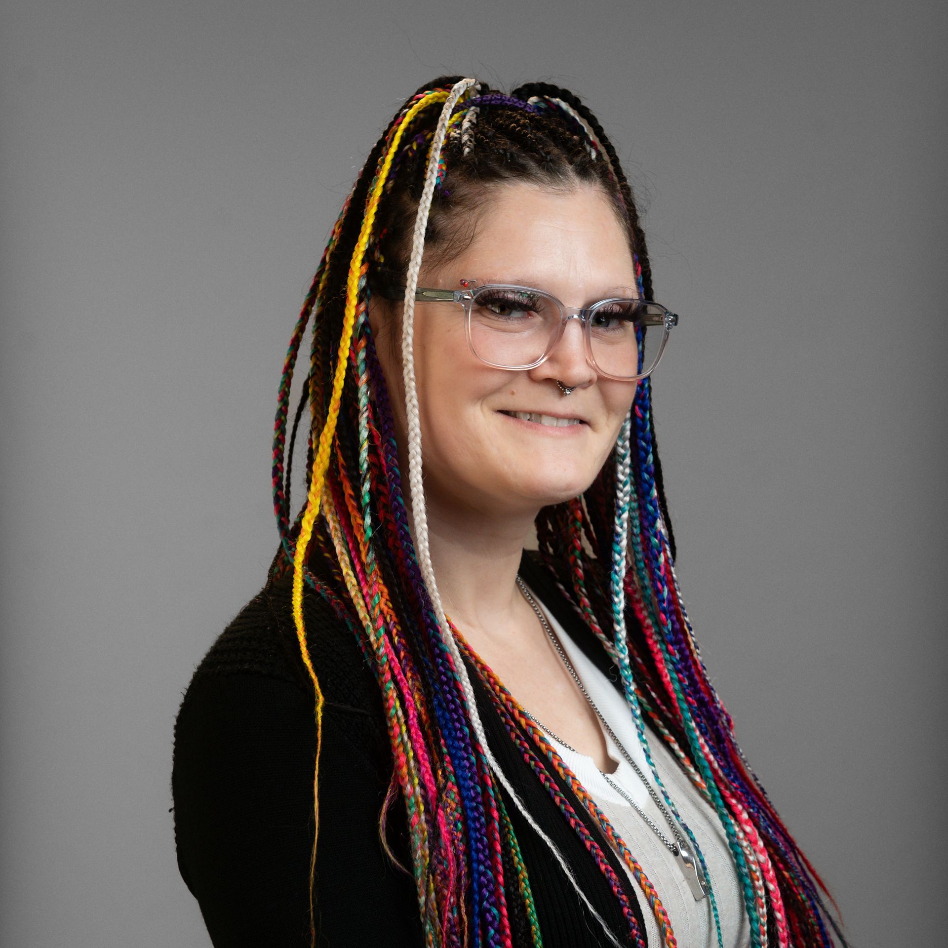 Woman with colorful braided hair and glasses smiles, wearing a black blazer.