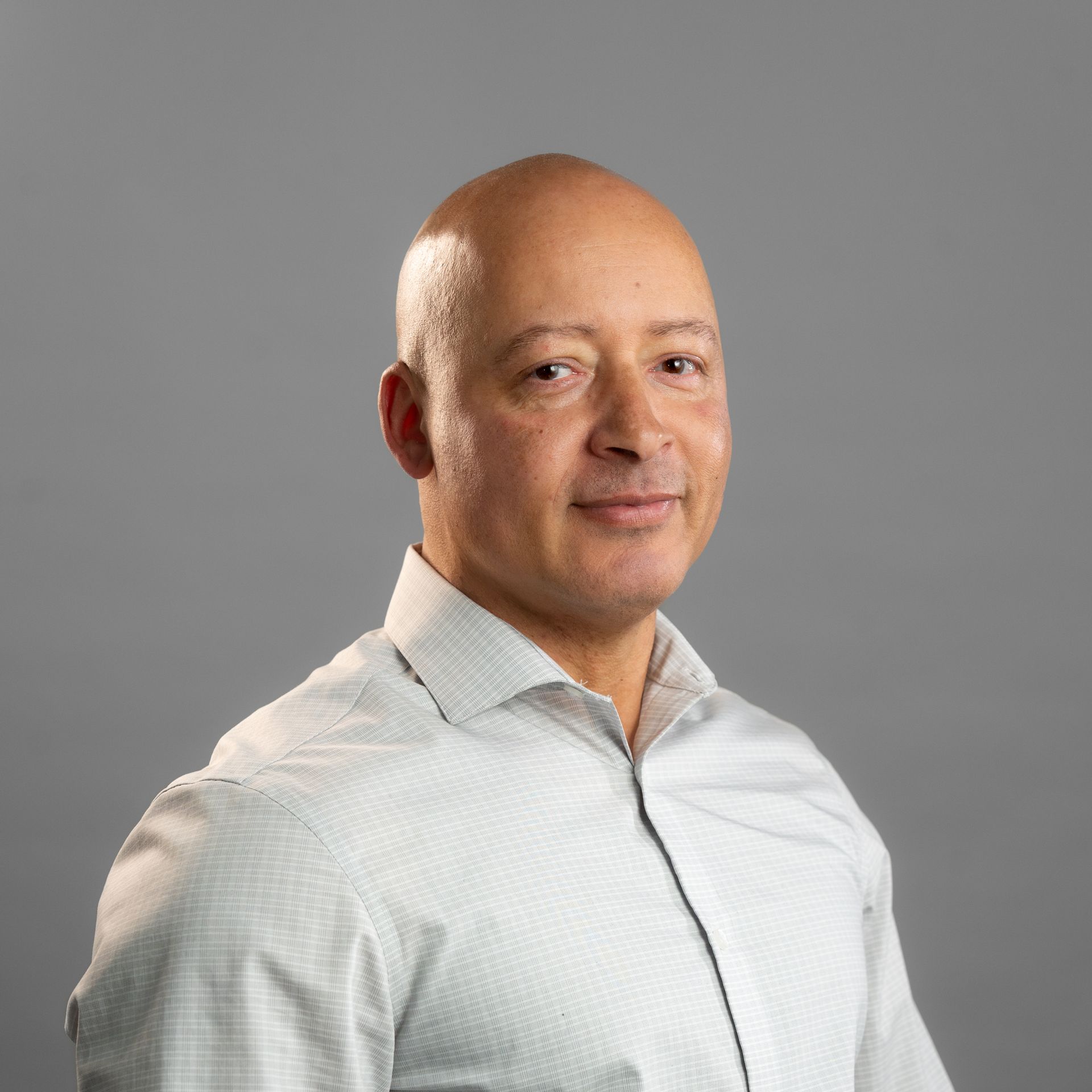 A bald man in a white shirt is standing in front of a gray background.