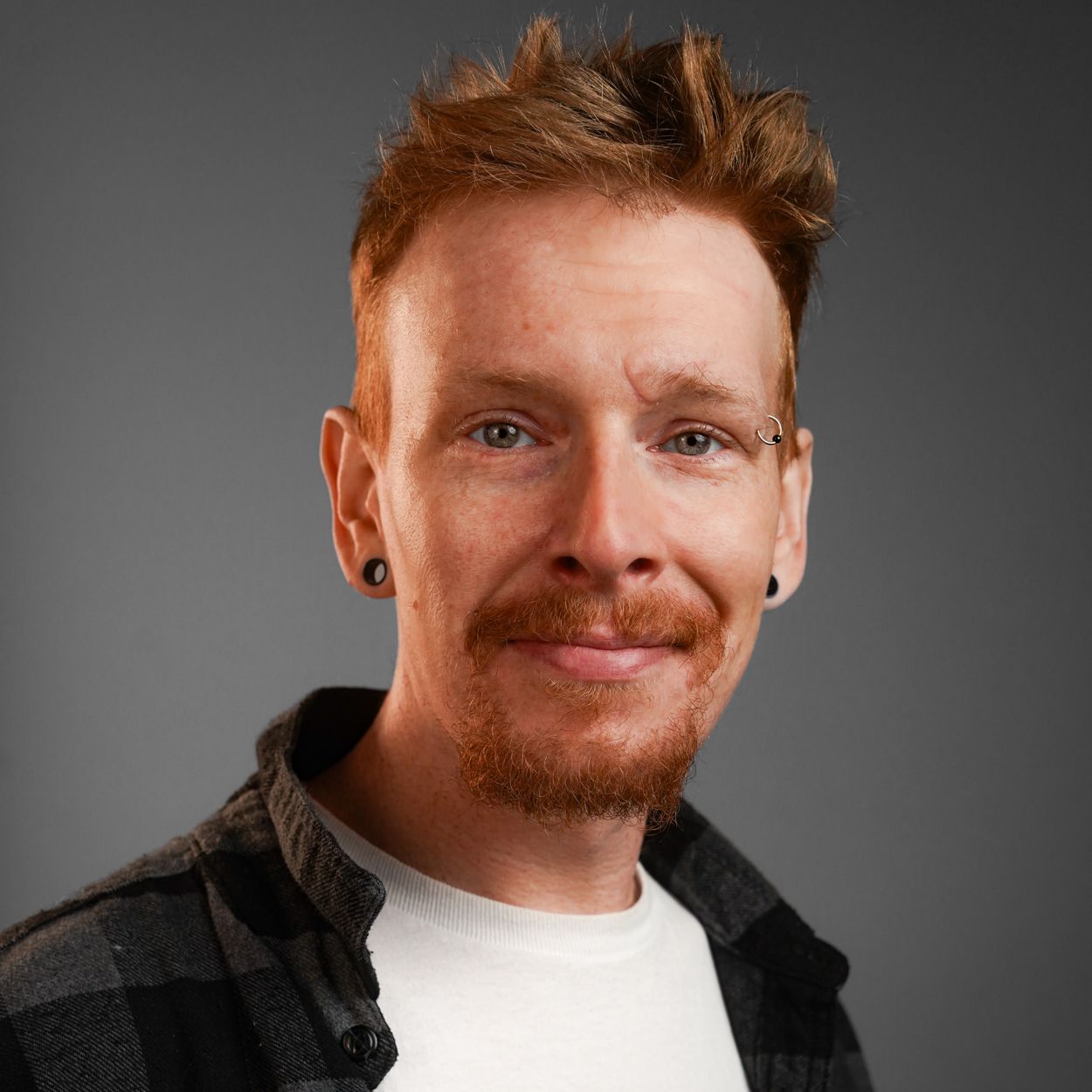 A man with red hair and a beard is wearing a plaid shirt and earrings.