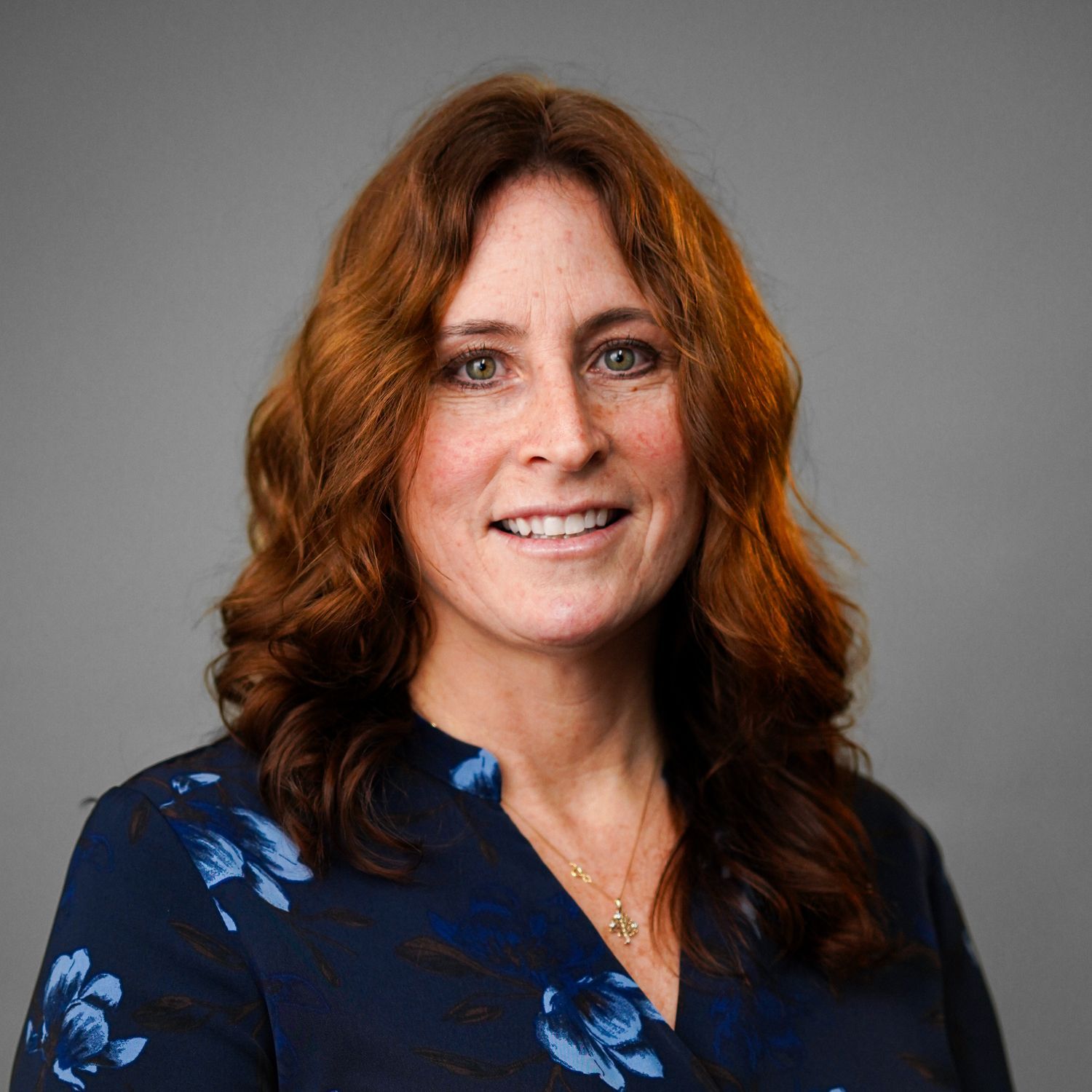 A woman with red hair is wearing a blue floral shirt and a necklace.