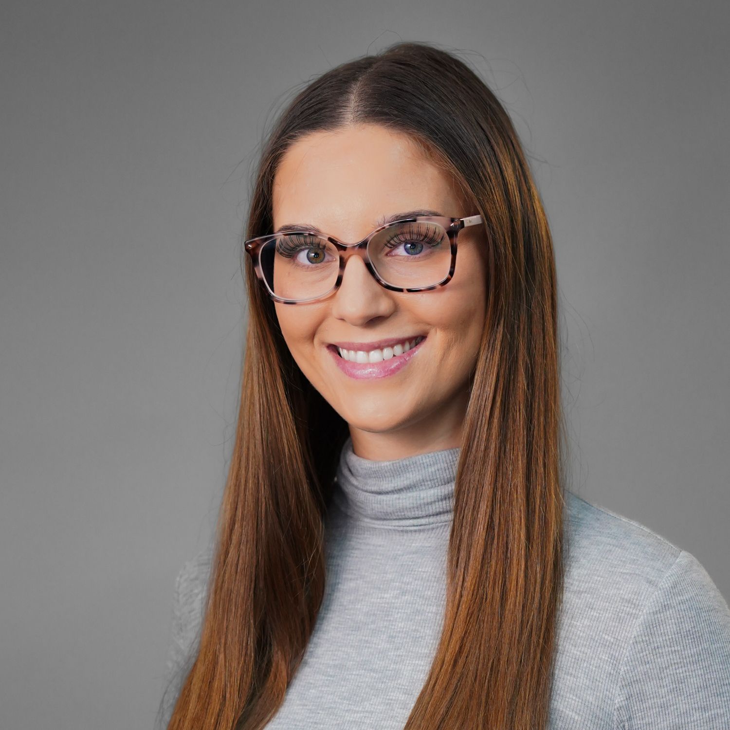 A woman wearing glasses and a grey sweater is smiling.