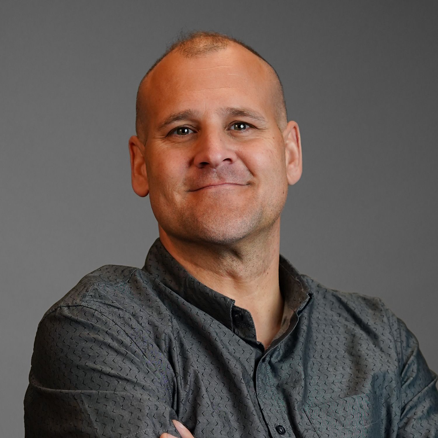 A man in a grey shirt is smiling with his arms crossed.