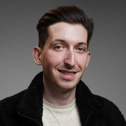 Young man with brown hair smiles, wearing a black fleece jacket, against a gray backdrop.