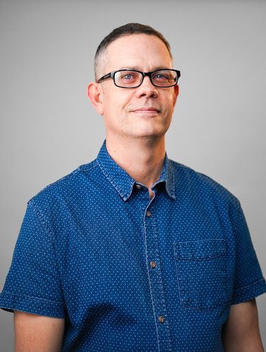 A man wearing glasses and a blue shirt is standing in front of a gray background.