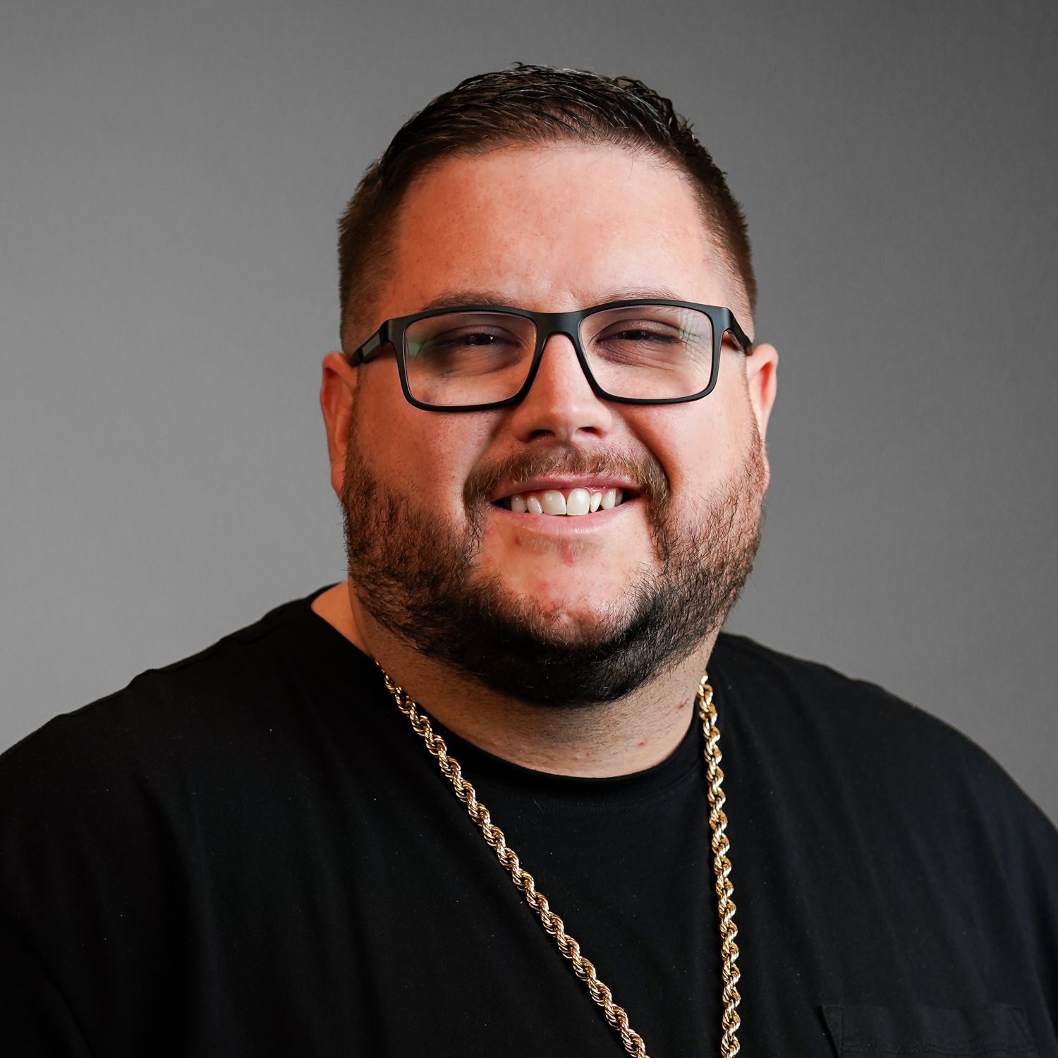 A man wearing glasses and a gold chain is smiling for the camera.