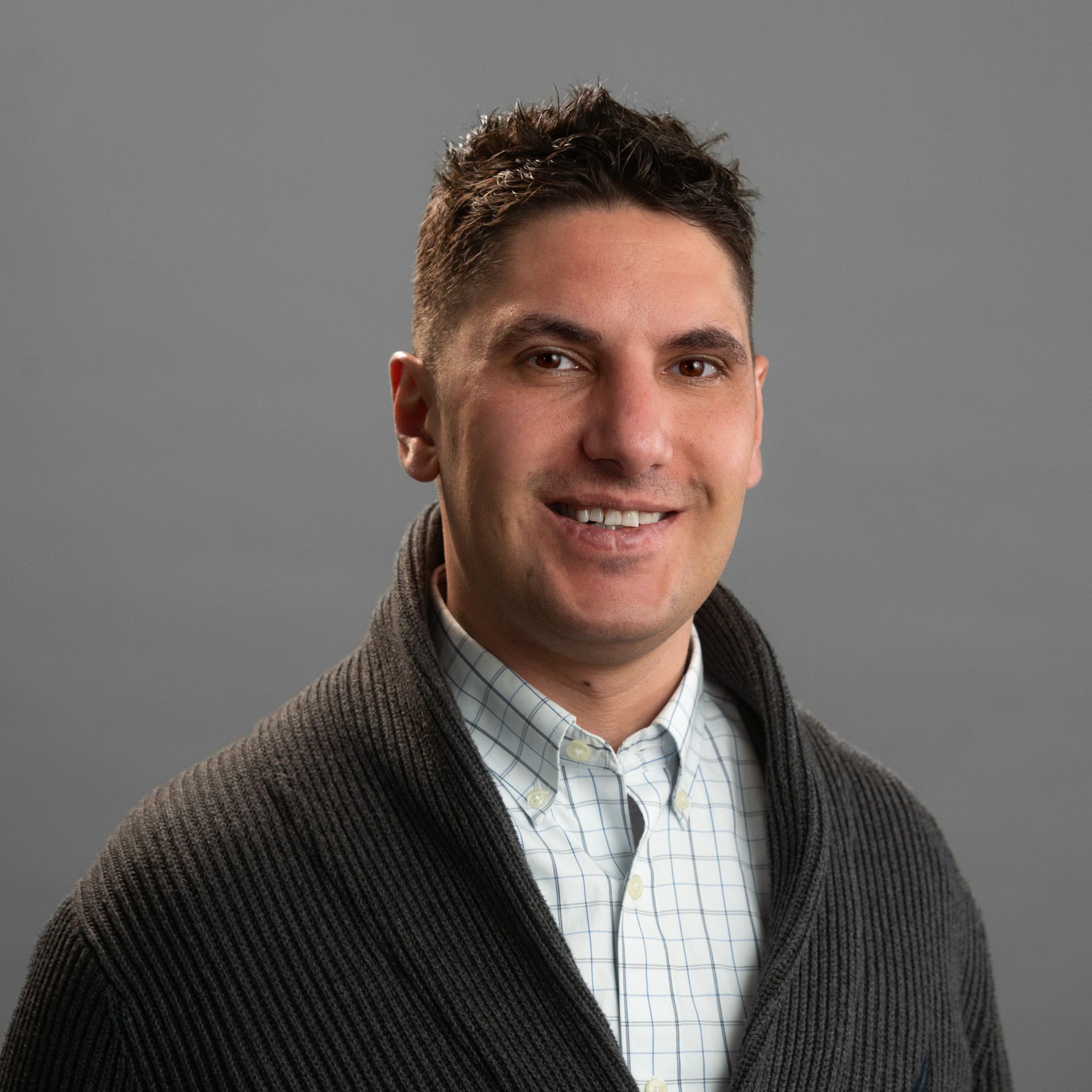 A man wearing a sweater and a plaid shirt is smiling for the camera.