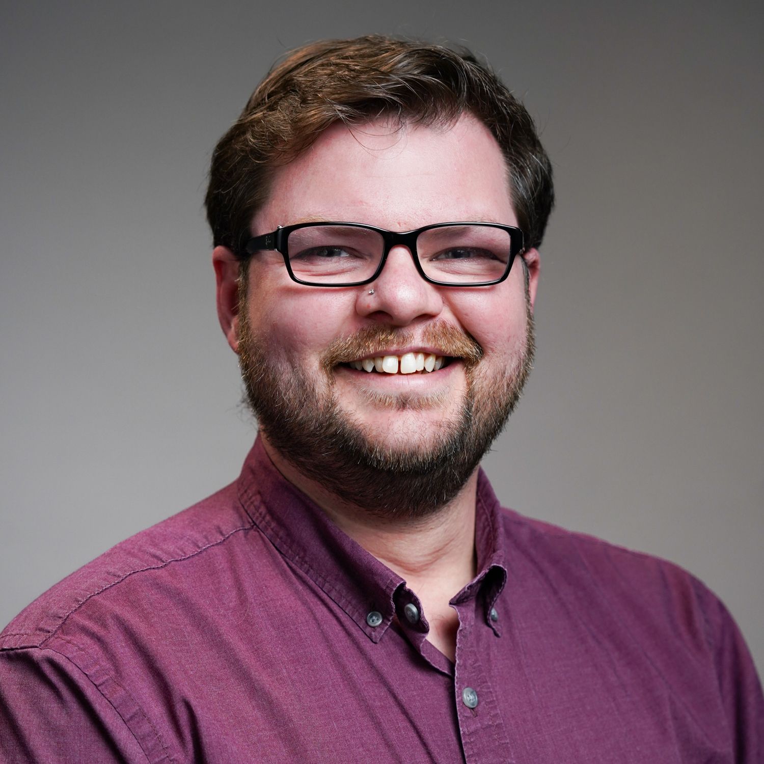 A man with a beard wearing glasses and a purple shirt is smiling.