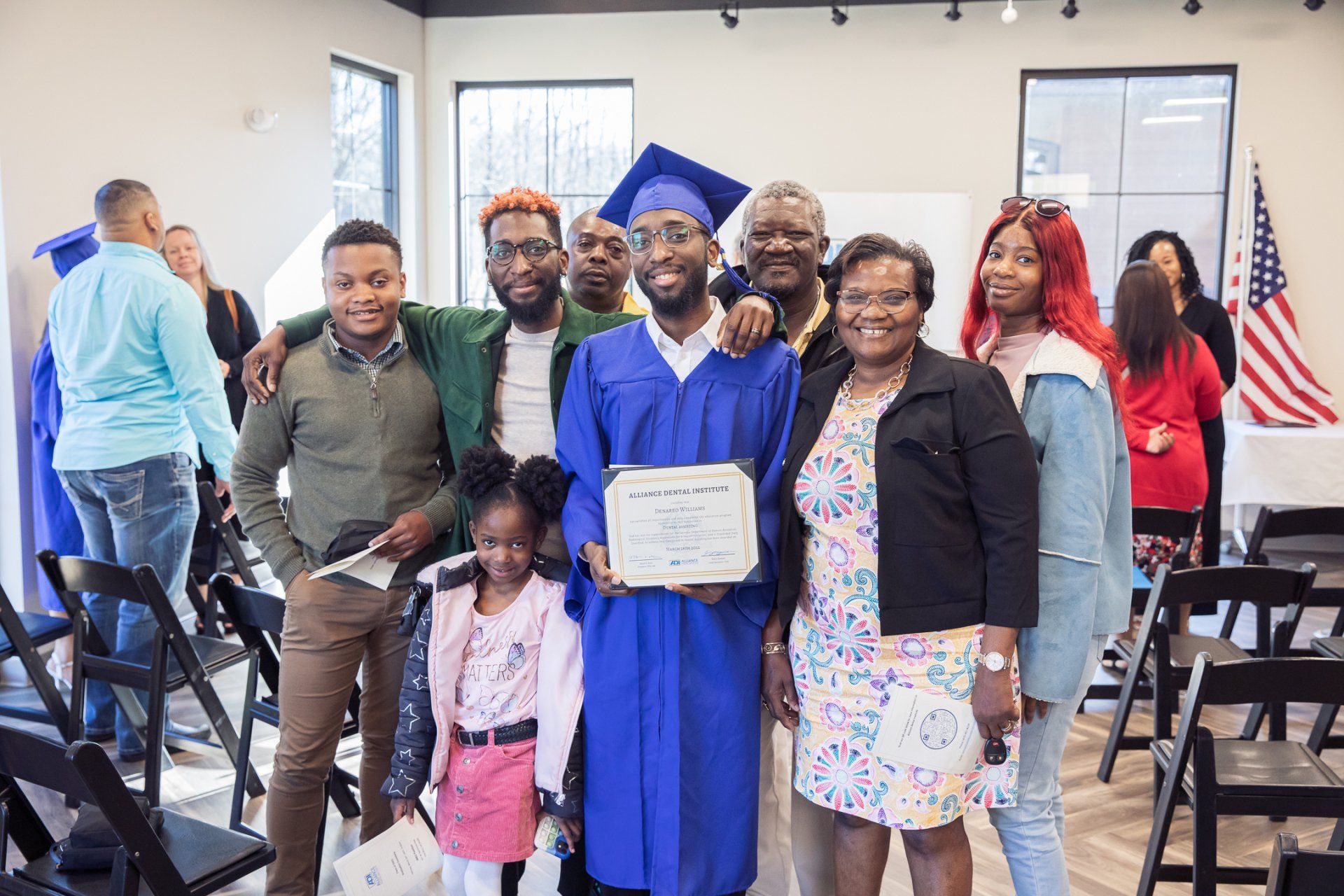 Family celebrates a graduate in blue cap and gown; indoors with an American flag in the background.