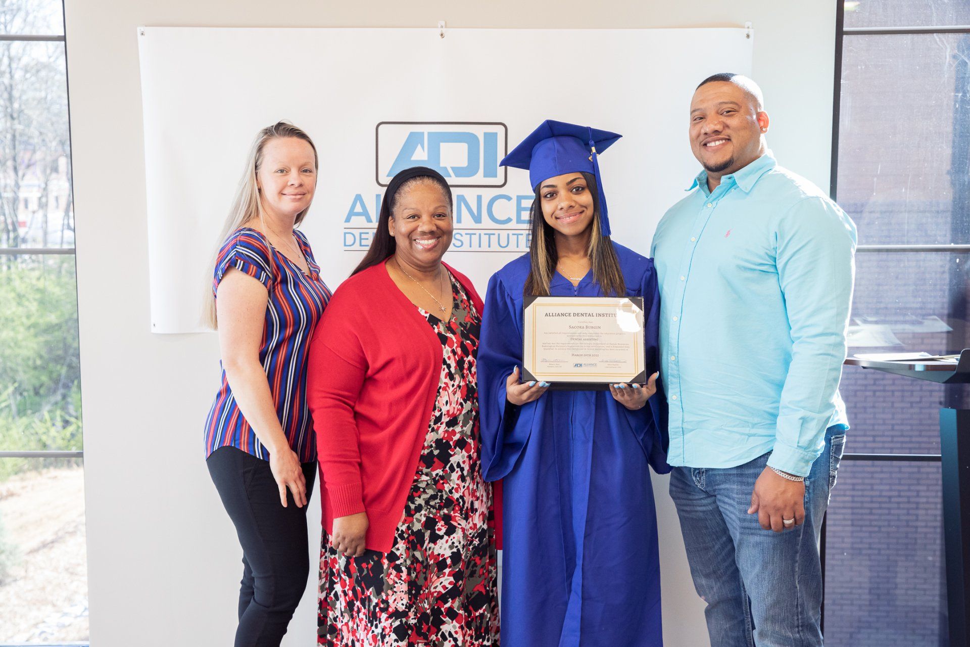 A graduate in blue cap and gown holds a diploma with family in a setting with a sign.