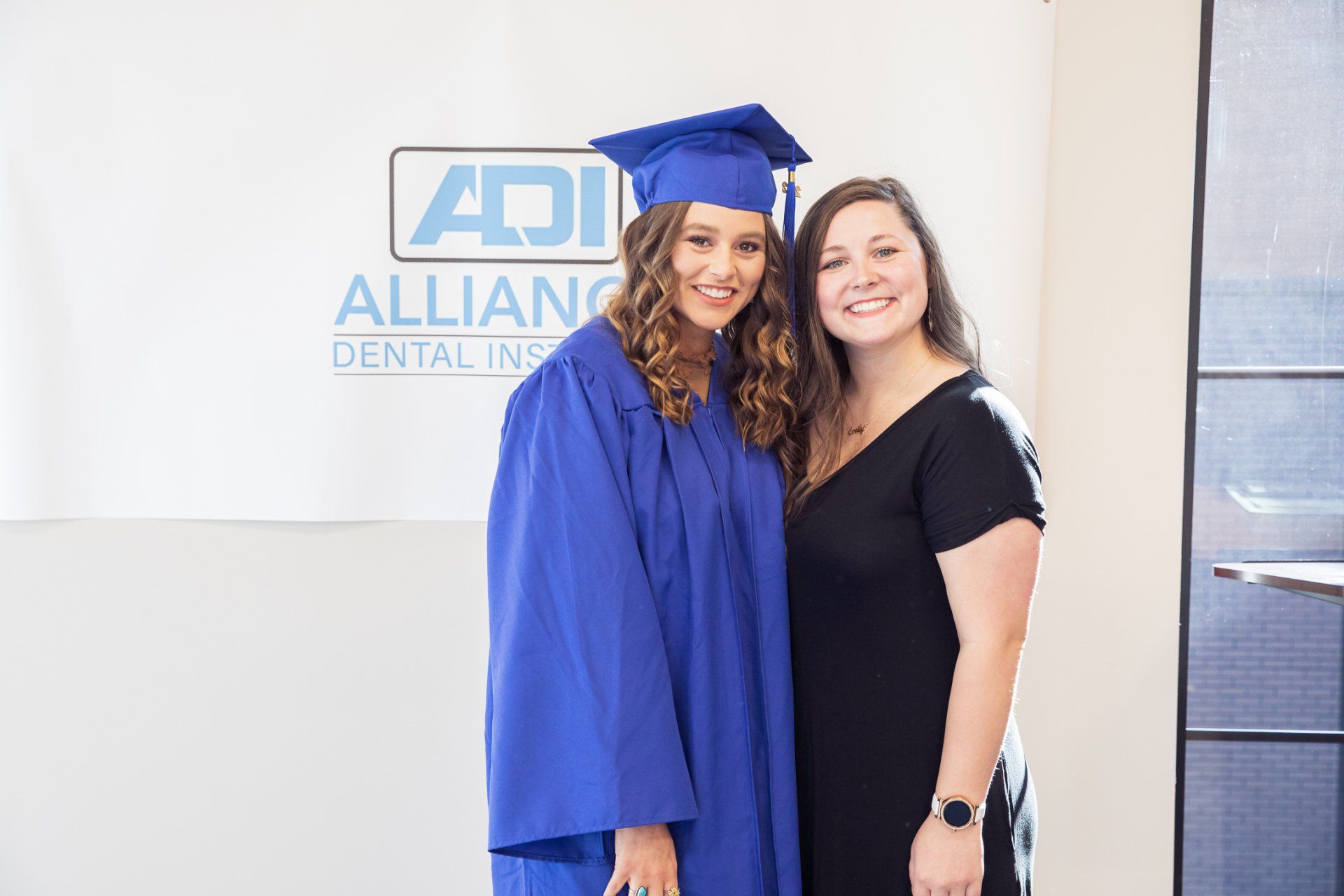 Woman in graduation gown poses with another woman in a dental school setting, smiling.