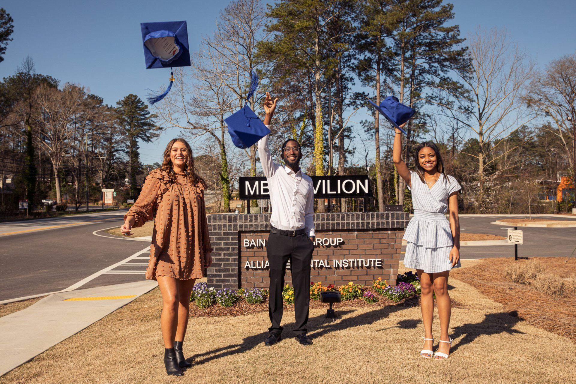 Three graduates in front of a pavilion throw their caps in the air, celebrating graduation.