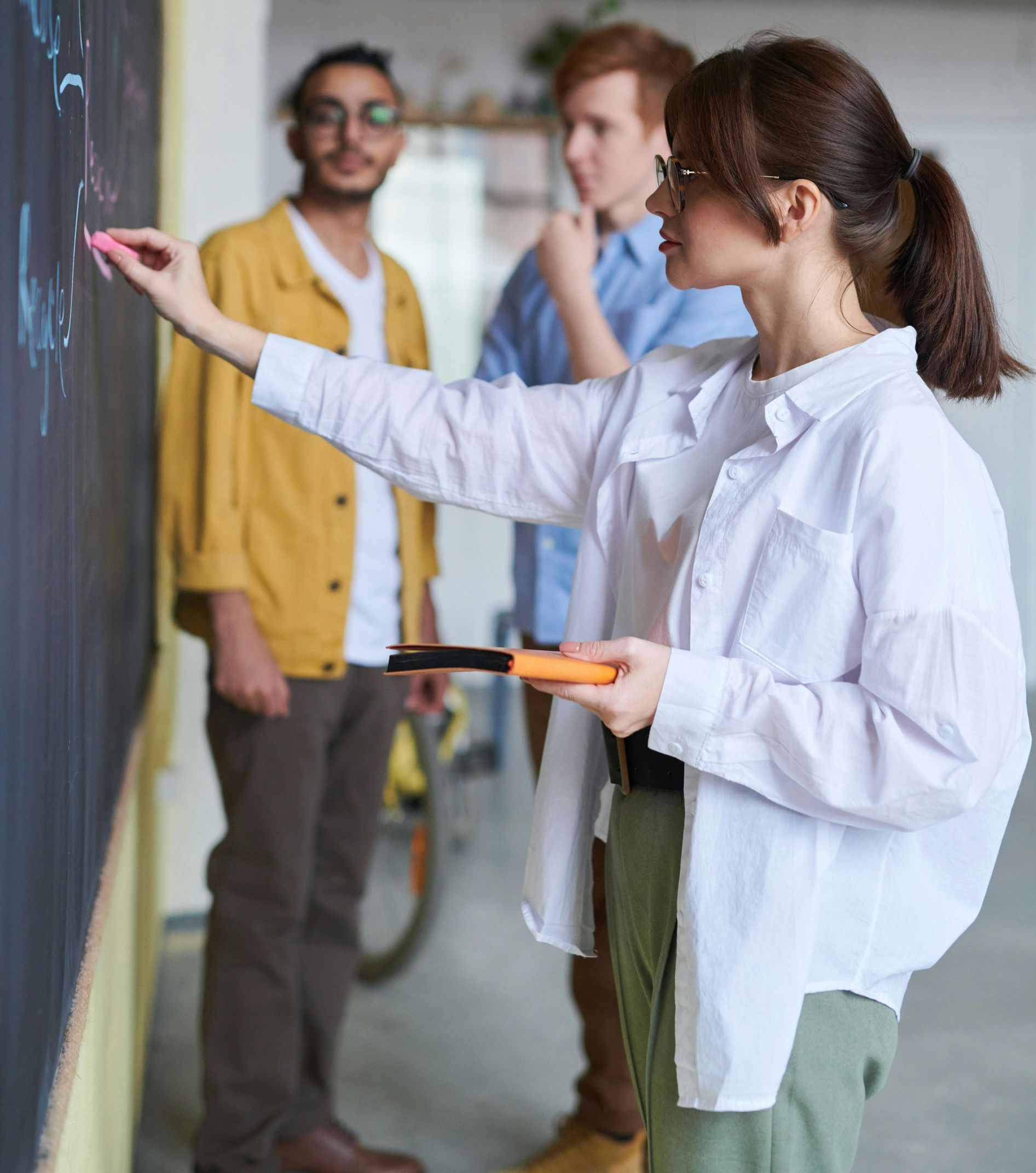 A woman is writing on a blackboard while two men look on.