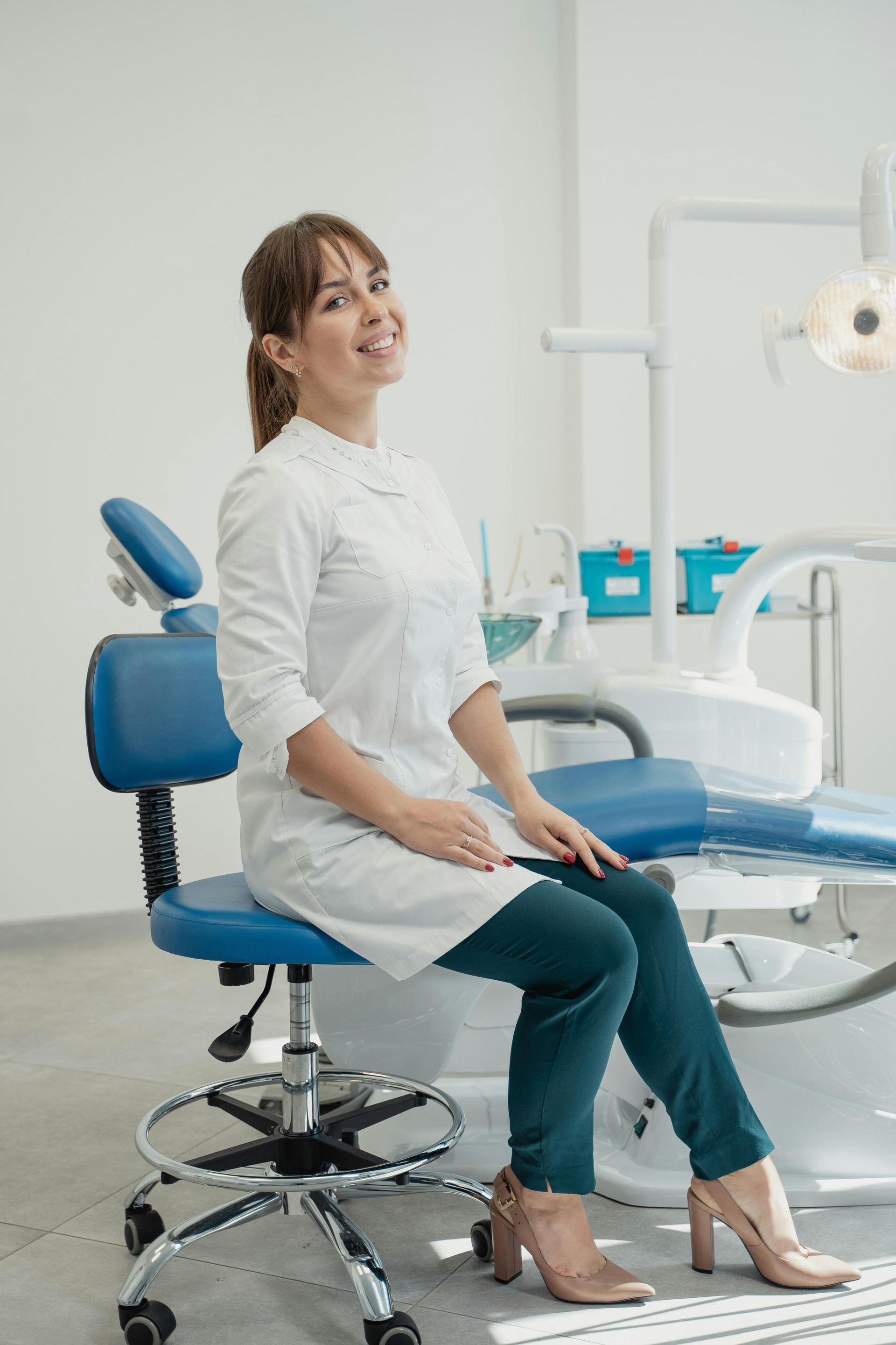 Woman in white coat smiles, sitting in dentist chair. Blue chair, dental equipment in the background.