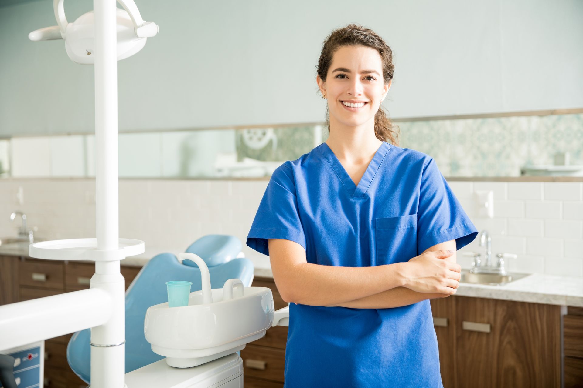 A smiling dental professional in a blue scrub top stands with arms crossed in a bright, modern dental office.