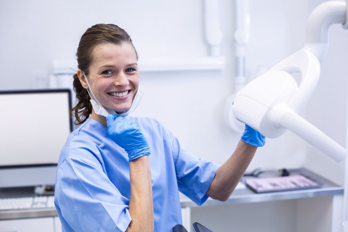 A dental assistant in blue scrubs smiling beside a dental light machine