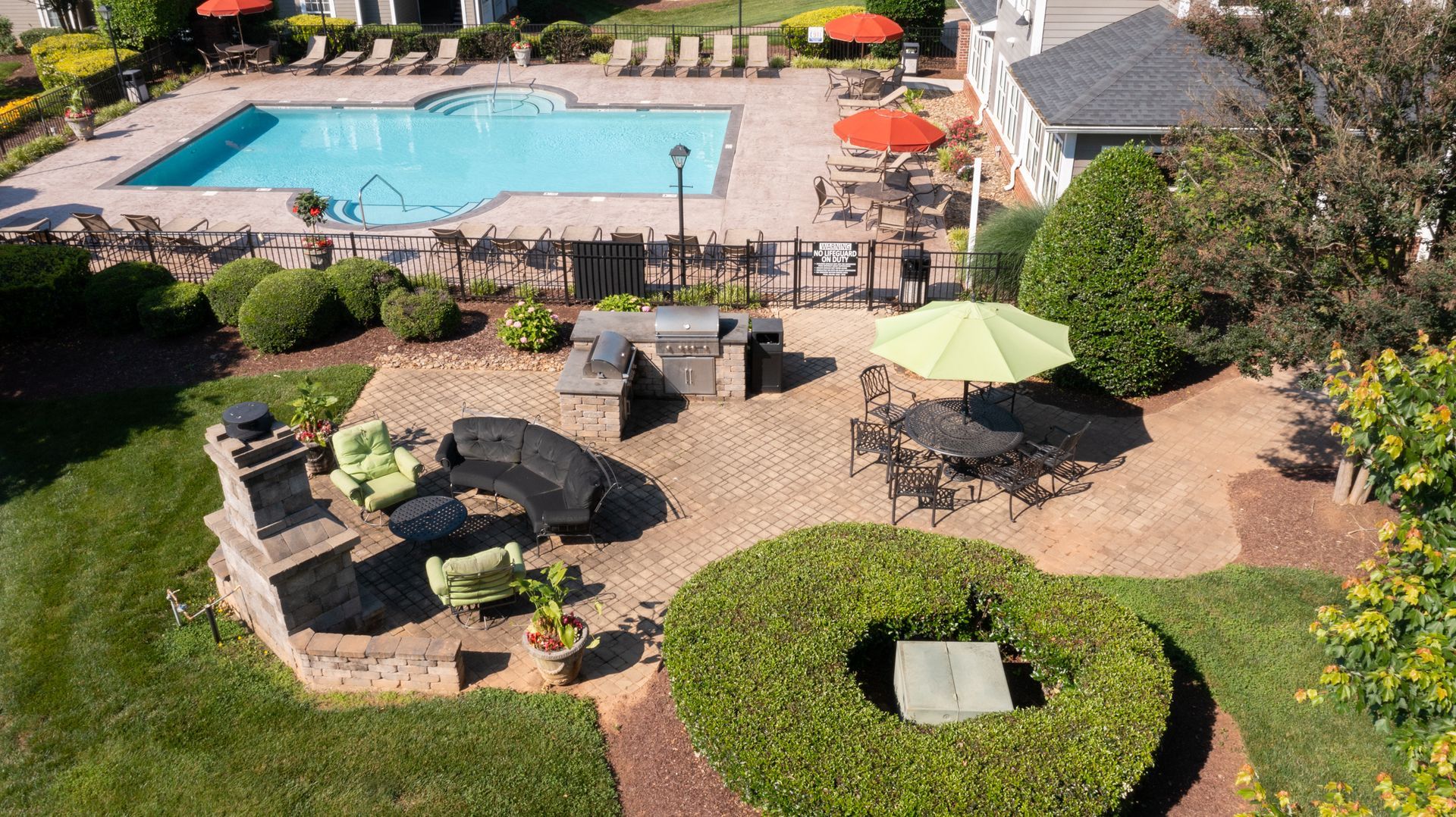 Poolside patio with grill and seating; pool, umbrellas, and lawn in background.
