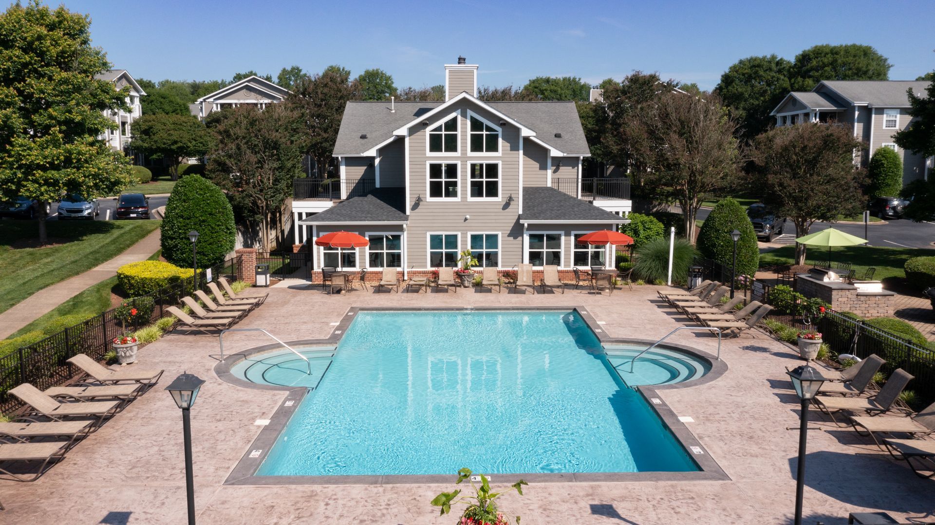 Pool and lounge chairs in front of a gray building with a red awning. Trees and blue sky surround the area.