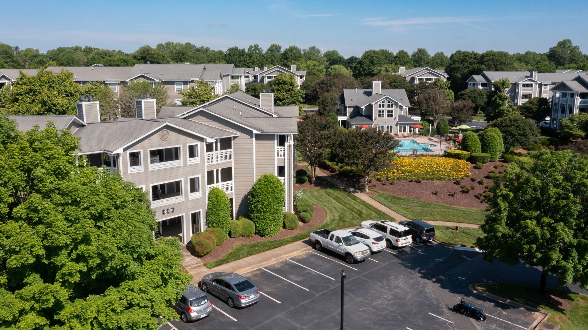 Apartment complex with gray roofs, parking lot, and swimming pool surrounded by trees and green lawns.