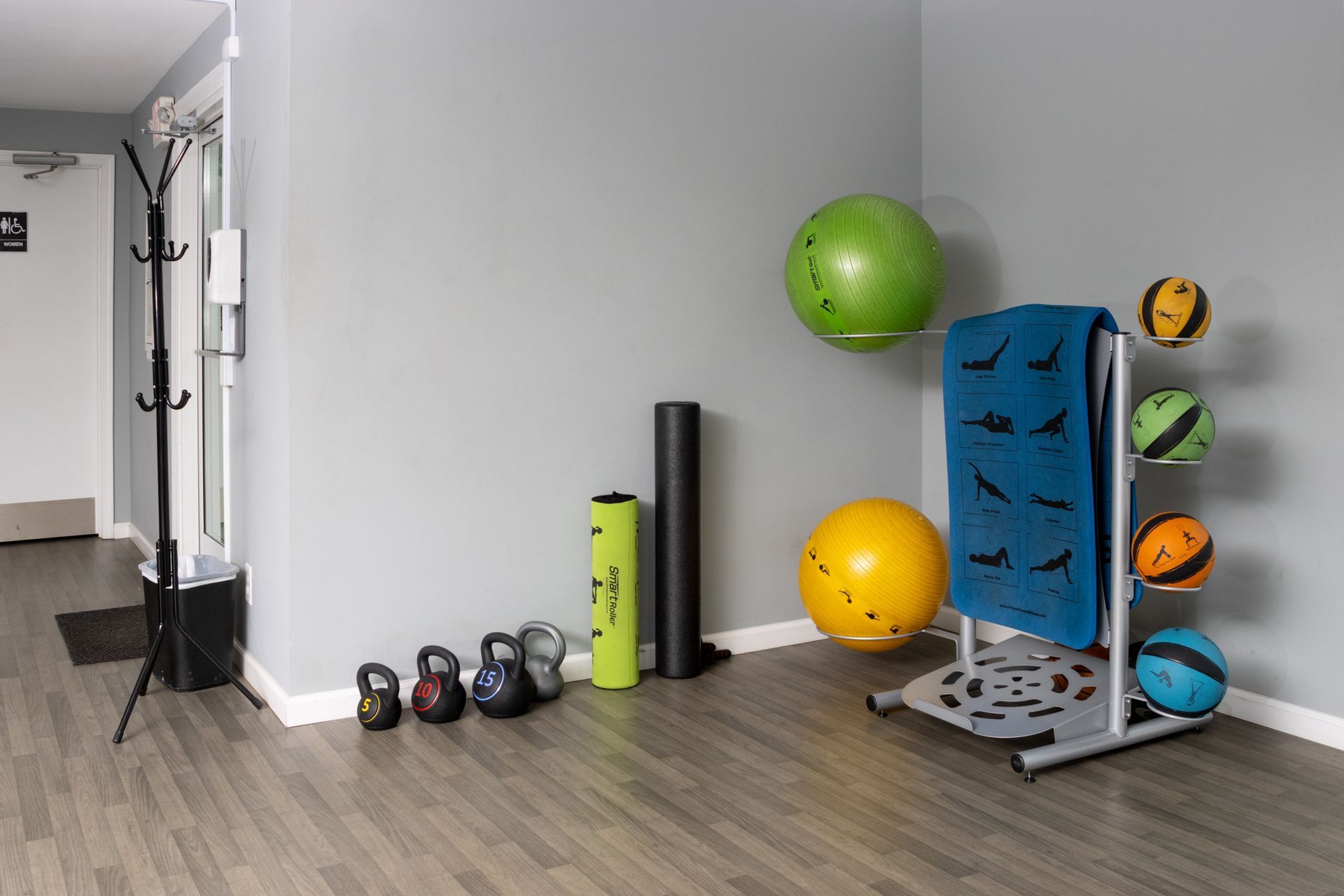 Gym corner with exercise equipment, including medicine balls, kettlebells, and a yoga mat, against gray walls.