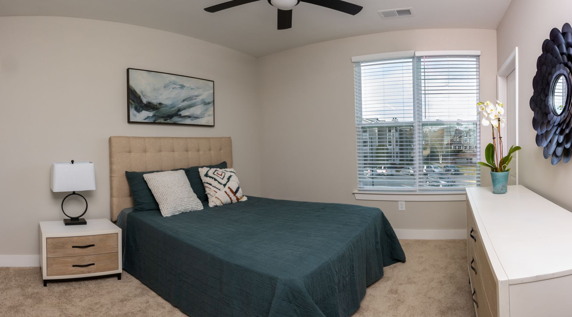 Bedroom with teal bedspread, beige walls, and a window. A dresser and nightstand are visible.