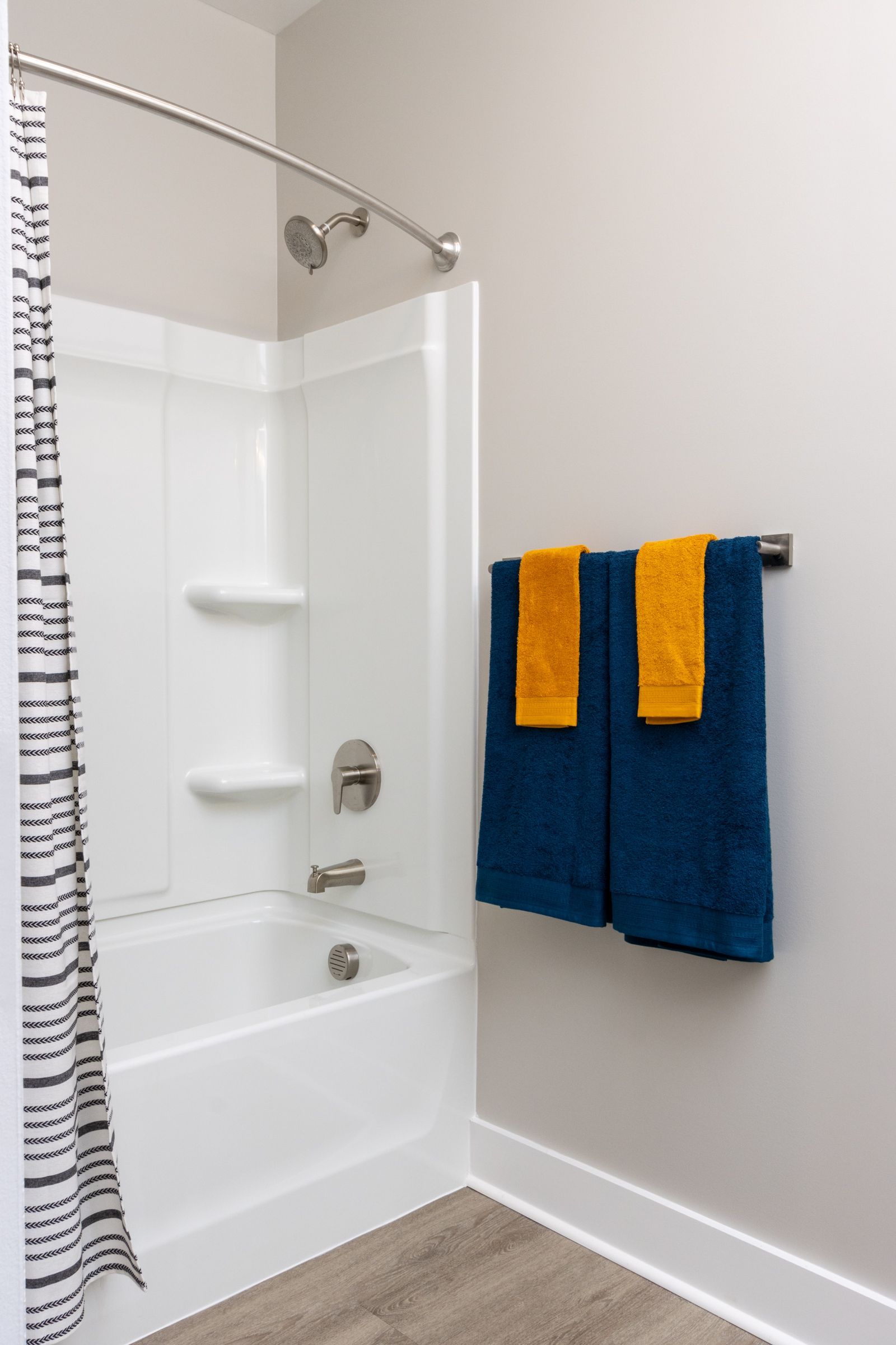 White shower/tub with two shelves, blue and gold towels. Light grey walls, wooden floor.