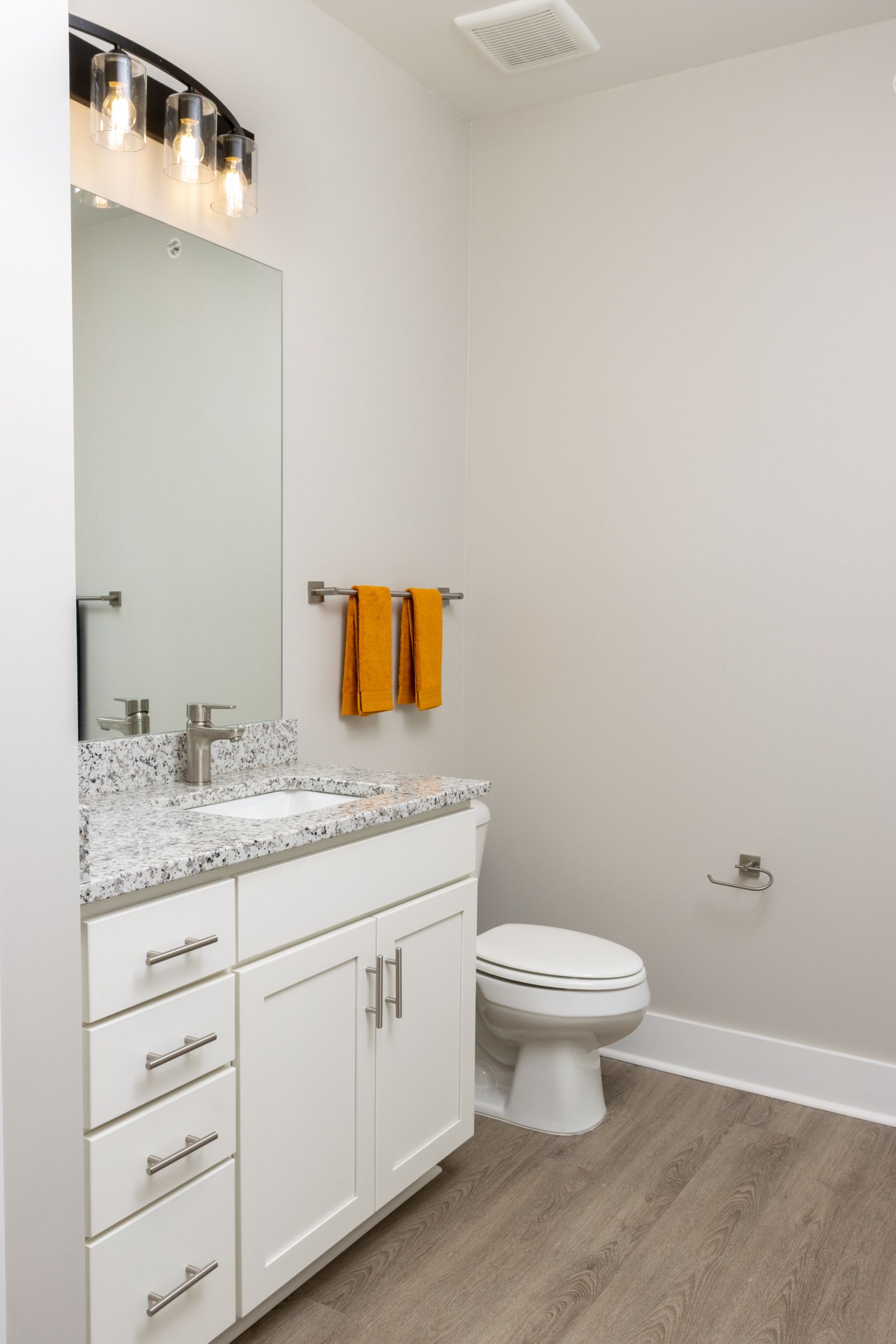 Bathroom with white vanity, toilet, and gray granite countertop. Orange towels hang on a silver bar.