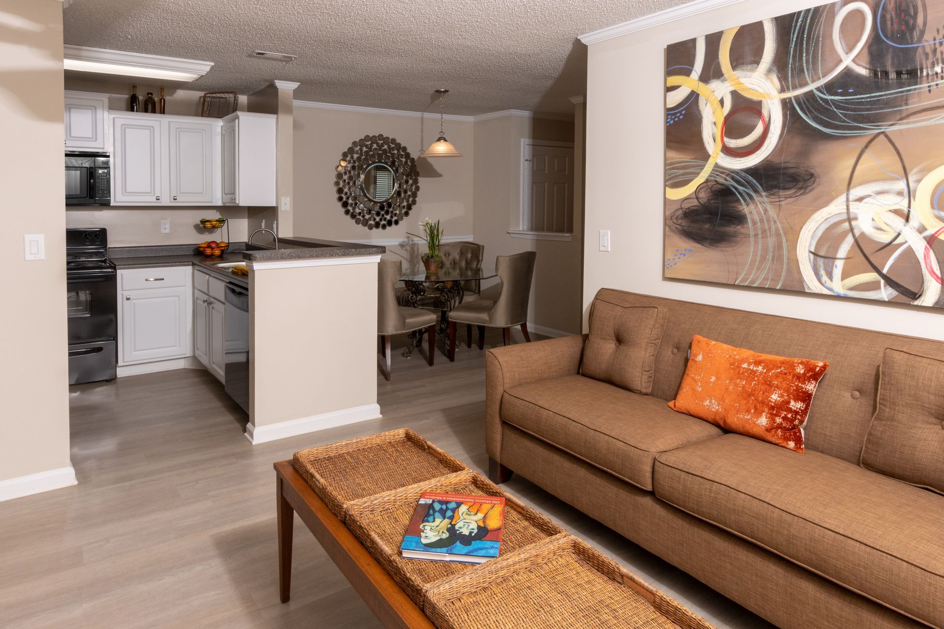 Living room with kitchen in background; brown sofa, coffee table, artwork.