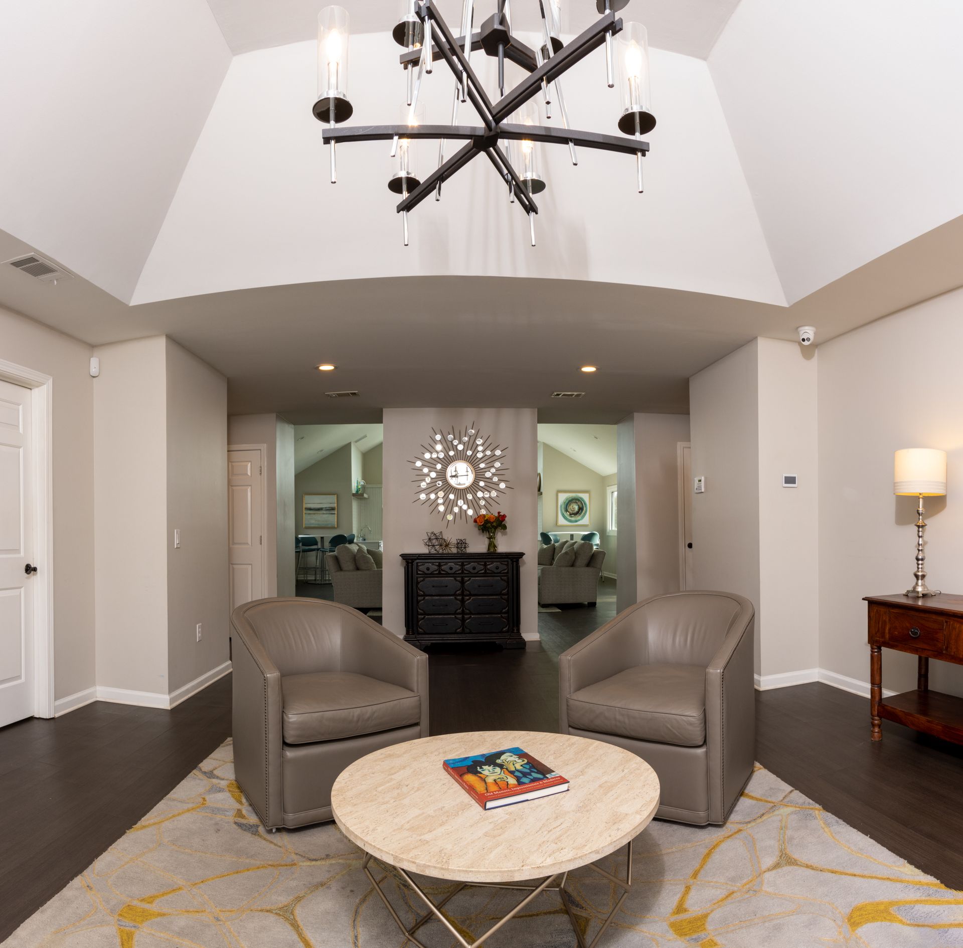 Living room with two gray chairs, a round table, and a chandelier. A doorway leads to another room.