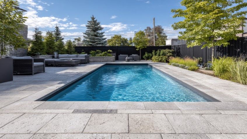 A rectangular swimming pool with blue water sits in a patio area surrounded by landscaping and a black privacy fence.
