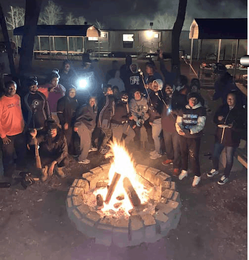 A group of people are standing around a fire pit at night.