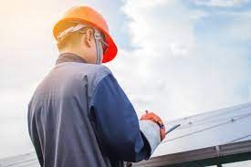 A man wearing a hard hat is writing on a clipboard in front of a roof.
