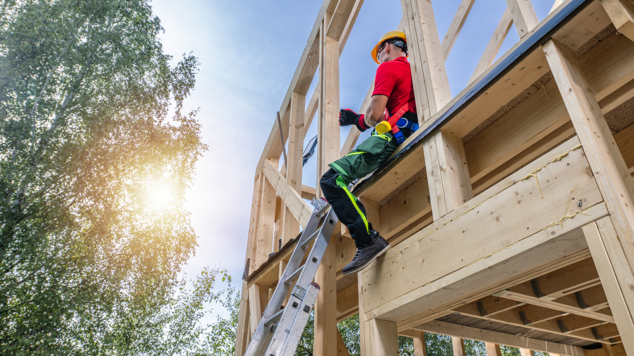 Construction worker on a wooden frame, wearing a hard hat, working under a sunny sky.