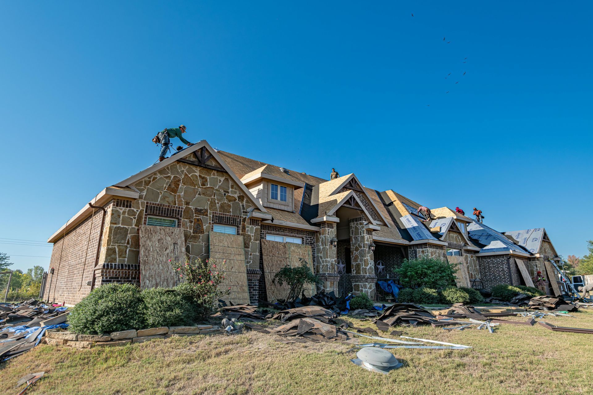 A person works on a roof. A house with stone siding under a clear blue sky.