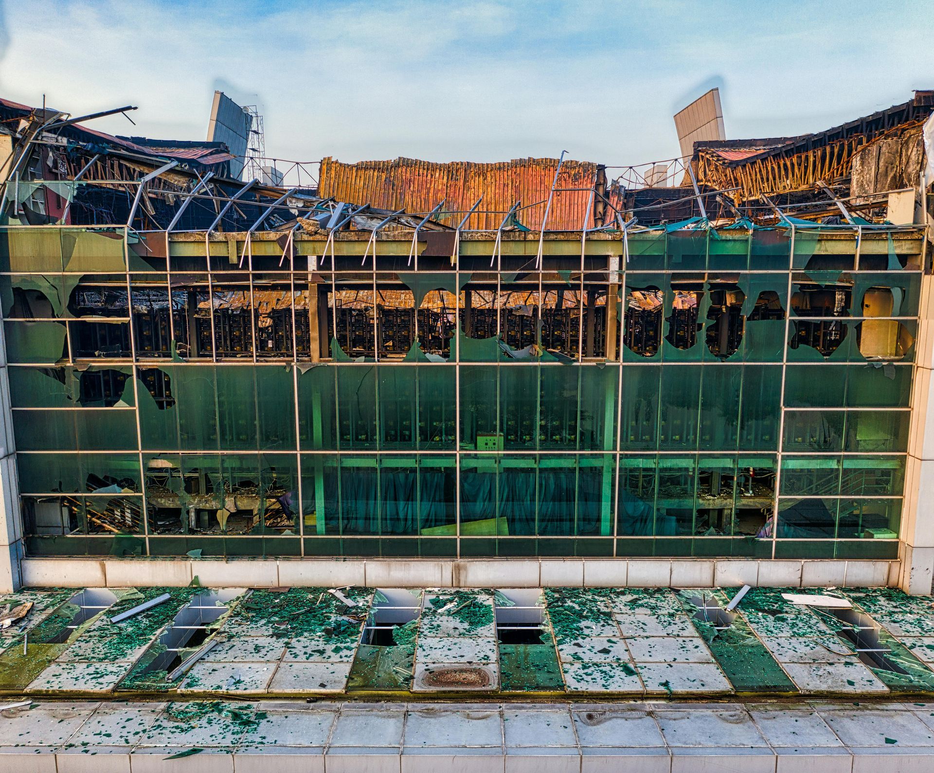 Damaged building facade; broken windows, scorched interior, debris. Green and blue color tones, clear sky.