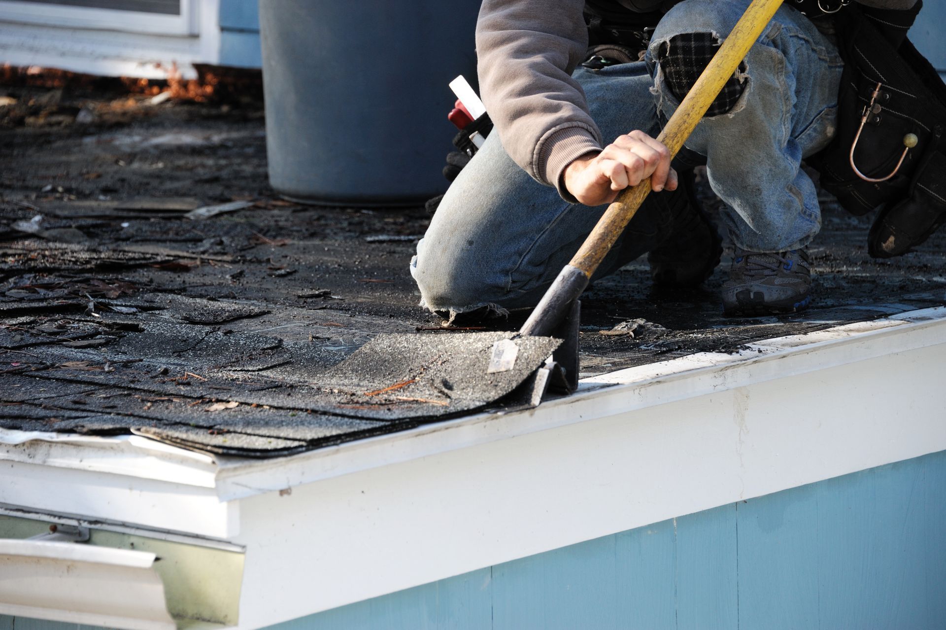 Roofer kneeling, removing old shingles from roof edge with shovel; blue house, sunny day.