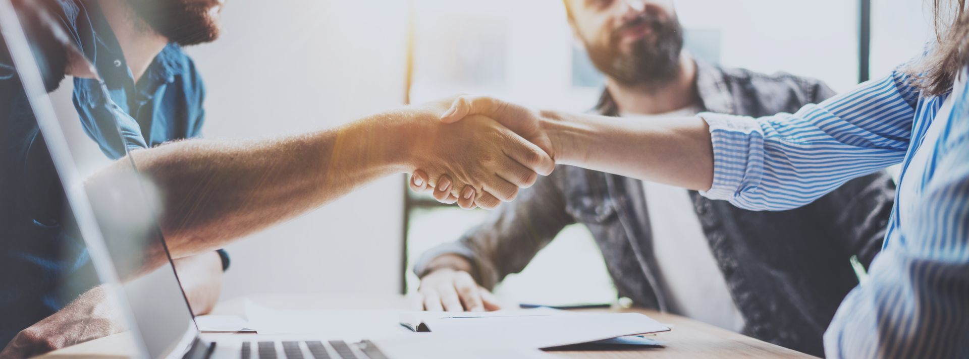 People shaking hands over a table with a laptop, possibly closing a deal in an office.