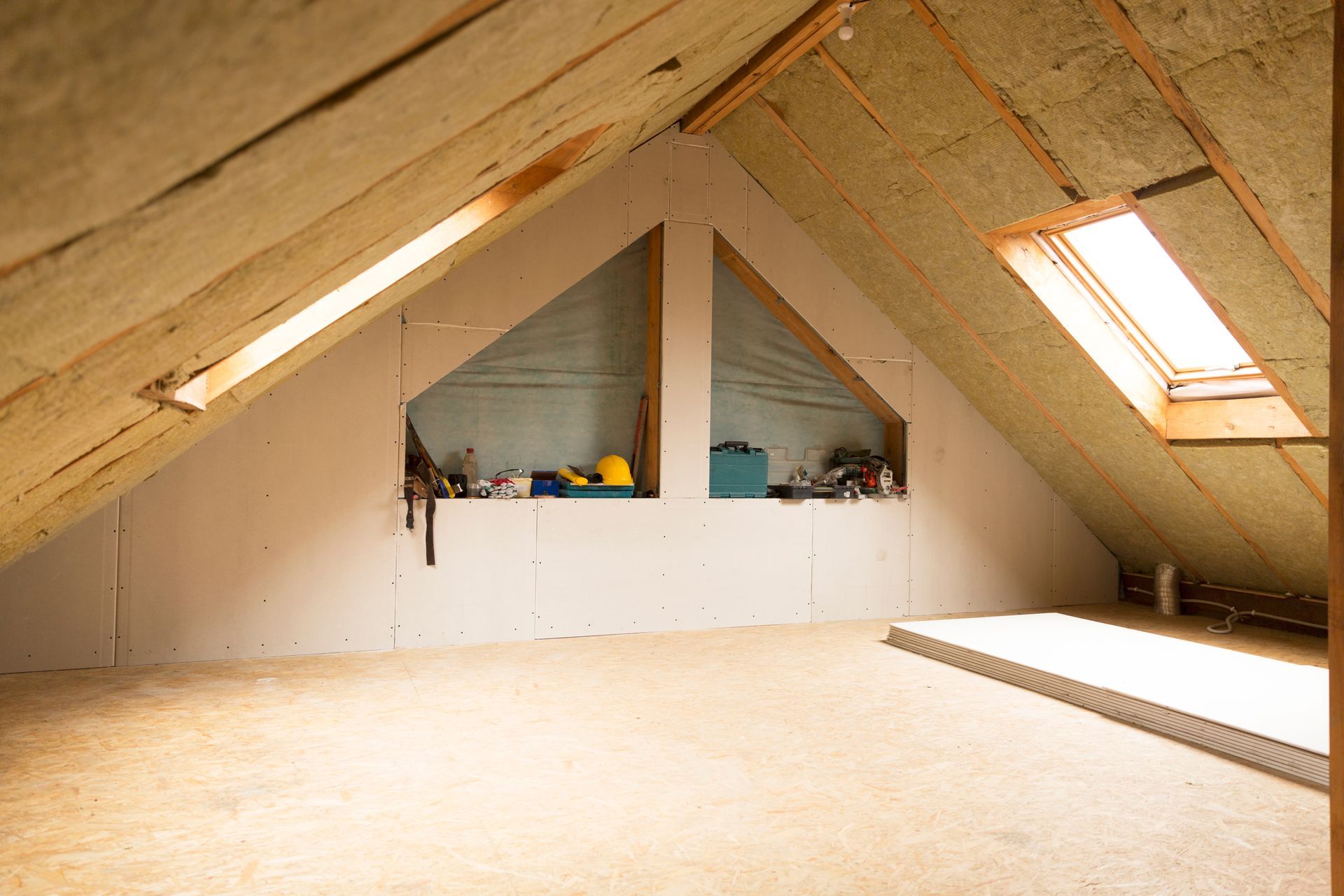 Attic interior with sloped ceiling, insulation, dormer windows, and a workspace with tools.
