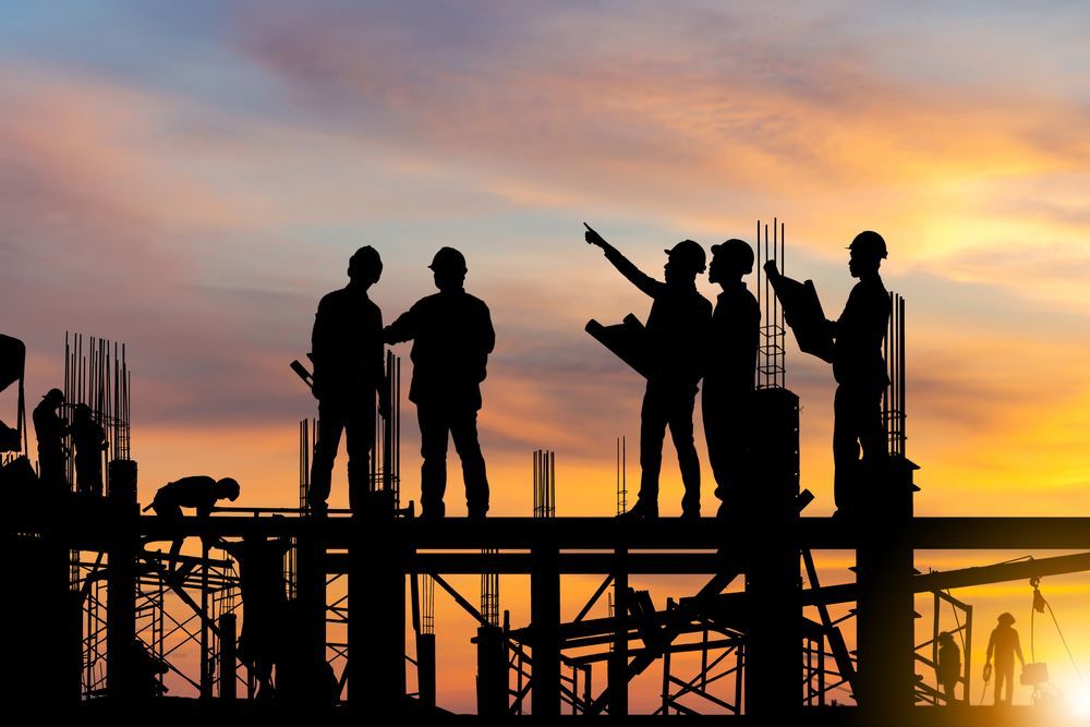 Silhouetted construction workers on a partially built structure, pointing at plans, sunset sky.