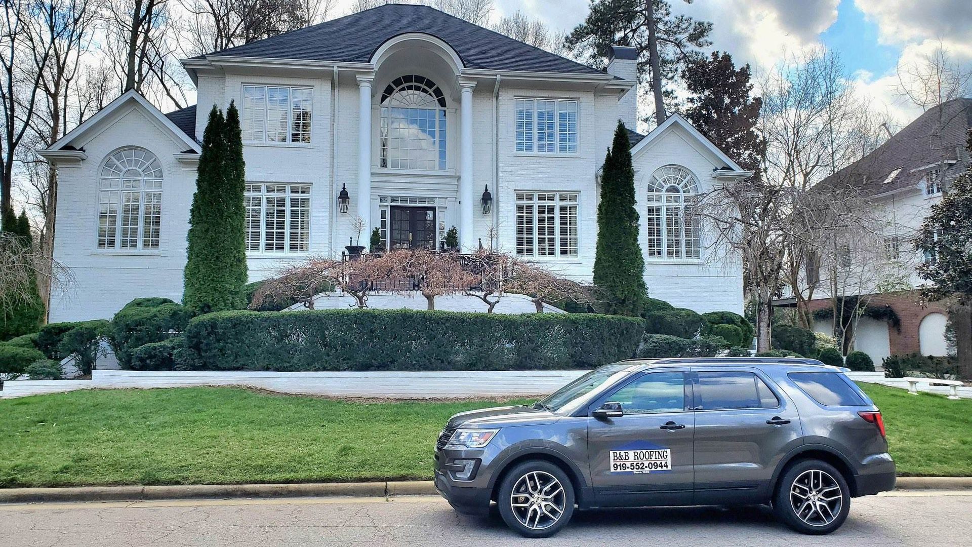 Grey SUV parked in front of a large white house with trimmed landscaping and trees.