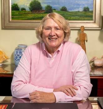 A woman in a pink shirt is sitting at a desk in front of a painting.