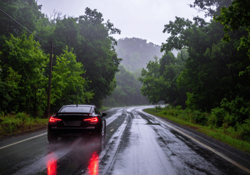 Car driving on a wet forest road in heavy rain with taillights reflecting on the pavement.