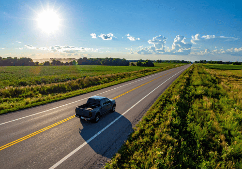 Pickup truck driving down a rural highway through open farmland under bright sunshine and blue skies.