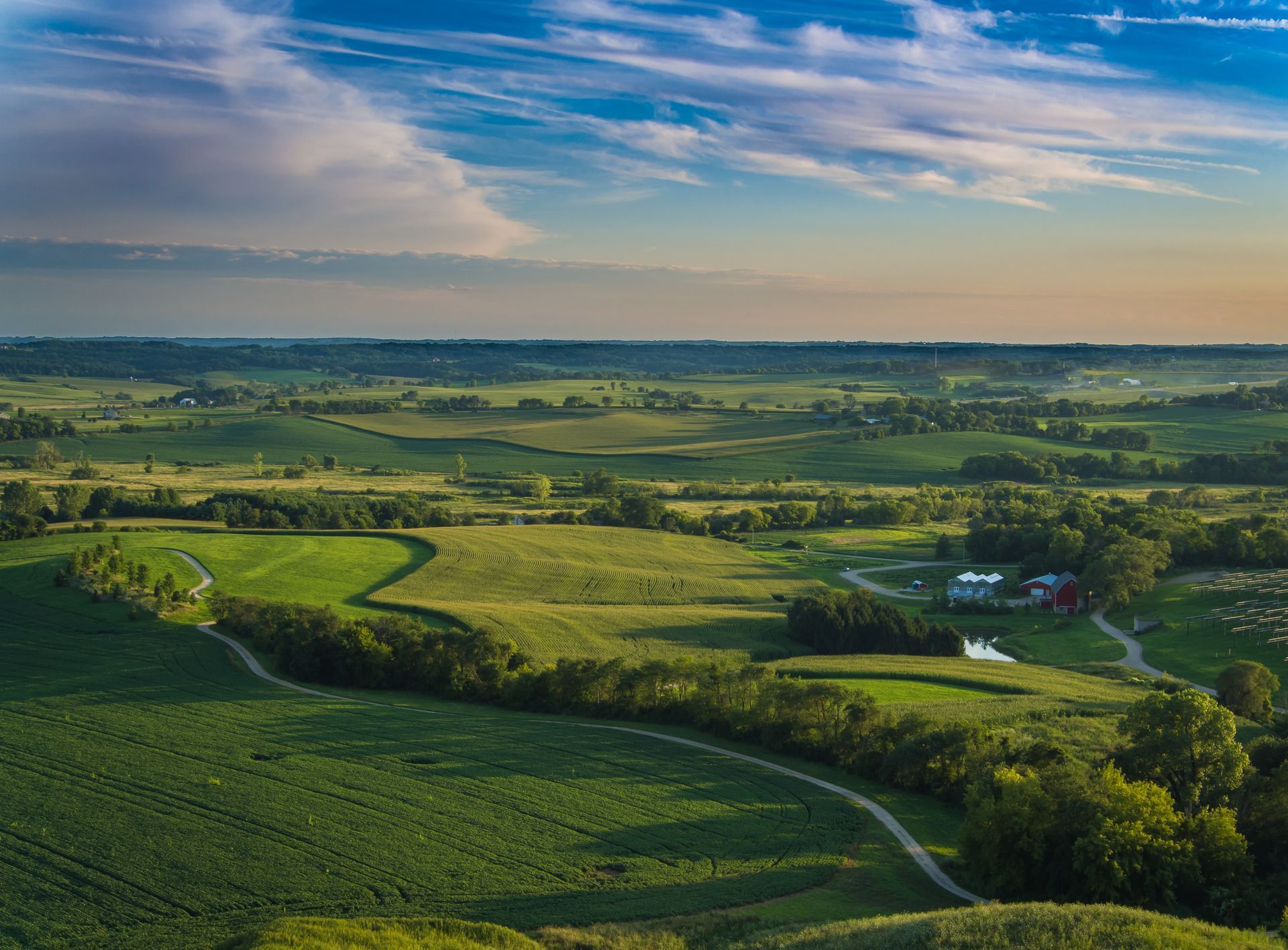 Lush green rolling hills under a soft, golden-hour sky with a winding path leading to a small rural farmstead.
