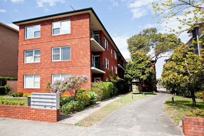 A three-story red brick apartment building with white window frames and a driveway leading to the back.