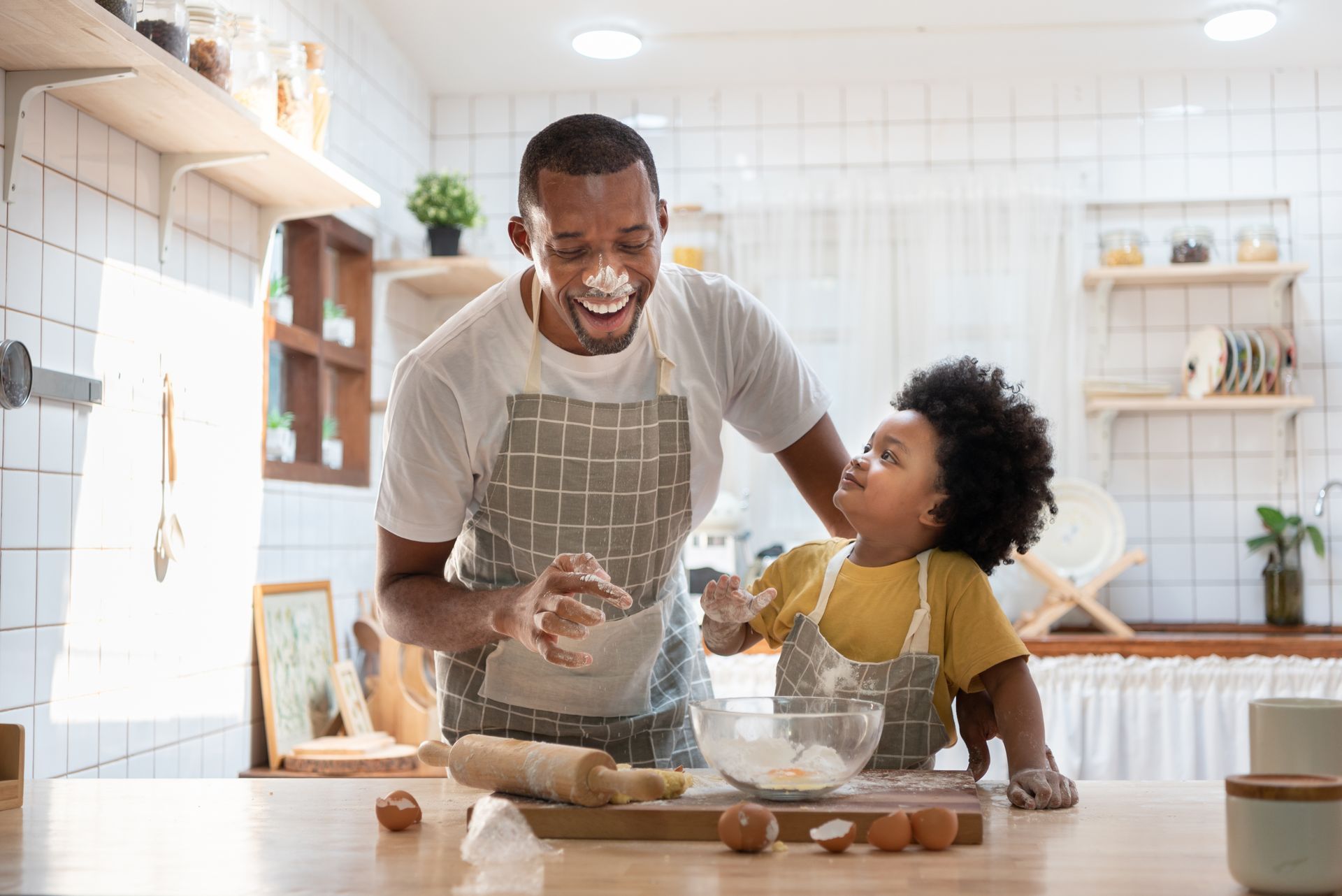 A parent and child wearing aprons bake in a kitchen, laughing together with flour on their faces and a bowl of dough.
